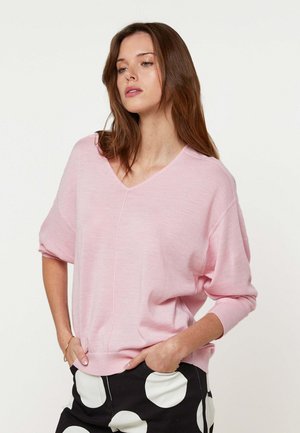 Vrouw met lang bruin haar die een lichtroze V-hals trui en zwarte broek met grote witte stippen draagt, handen in de zakken.