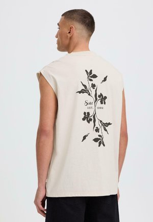 Homme portant un débardeur blanc cassé avec un motif floral noir et le texte « Solid EST. 1999 » au dos, assorti à un short noir.