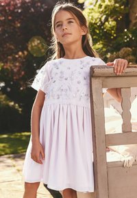Rochie albă cu mâneci scurte, din material transparent, decorată cu broderie florală și paiete. Fustă evazată cu pliseuri și decolteu rotund.