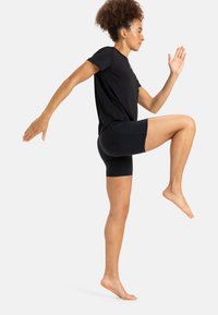 Sort atletisk skjorte og shorts. Skjorten er løstsiddende, og shortsene sidder tæt. Personen er barfodet med det ene ben løftet.