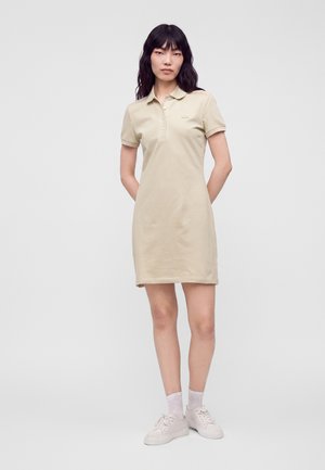 Une femme présente une robe polo beige à manches courtes, des chaussettes blanches et des baskets sur un fond clair et uni.
