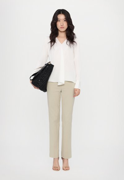 Blusa blanca de manga larga con escote en V, pantalones beige a medida y un bolso negro de tejido. La modelo está de pie con un fondo neutro y sandalias.
