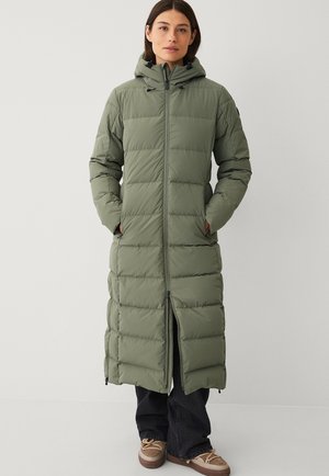 Lang, polstret frakke i olivengrøn med hætte, med lodret quiltet design, for-lynlås og sidelommer. Mørke jeans og brune sko synlige.