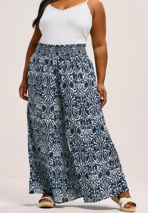 Donna che indossa pantaloni a gamba larga con motivo navy e bianco, canottiera bianca con bordo in pizzo e sandali con zeppa bianchi aperti in punta.