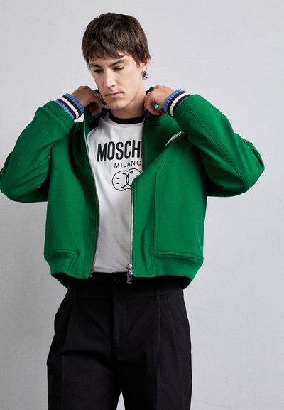 Zelená mikina na zips s rebrovanými manžetami, v kombinácii s bielym tričkom s potlačou. Čierne nohavice dokončujú celkový dojem.