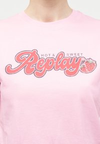 Άτομο που φοράει ανοιχτό ροζ πουκάμισο με το κείμενο "Hot & Sweet Replay" και δύο φράουλες τυπωμένες σε ροζ και κόκκινο στο στήθος.