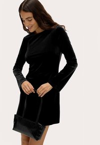 Zwarte fluwelen jurk met lange mouwen en een aansluitende pasvorm, gecombineerd met een kleine zwarte plisséhandtas met twee handvatten.