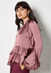 Rosa Satinbluse mit gerafften Schichten am Saum und an den Ärmeln. Hoher Kragen, leichter Stoff mit subtiler Sheen, taillierte Passform.