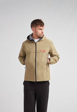 Beige jakke med hætte, sorte lynlåsdetailler og sidelommer. Har et "JACKIT" logo i rødt på brystet, glat tekstur.