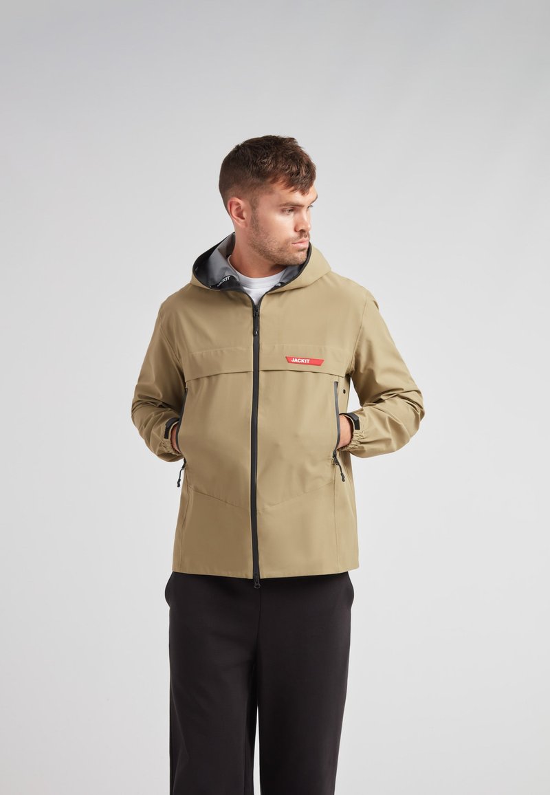 Beige jas met een capuchon, zwarte ritsdetails en zijzakken. Kenmerkt een "JACKIT" logo in het rood op de borst, met een gladde textuur.