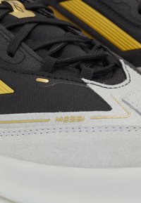 Sportschuhe mit einem schwarzen Textil-Obermaterial, grauen Wildlederelementen, goldenen Beschlägen und auffälligen Schnürsenkeln. "MESSI" ist an der Seite eingeprägt.