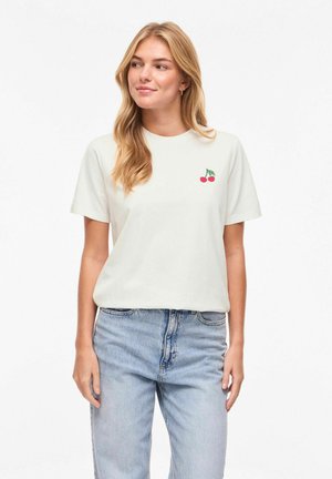 Femme blonde portant un t-shirt blanc avec un petit motif de cerise sur la poitrine et un jean taille haute bleu clair.