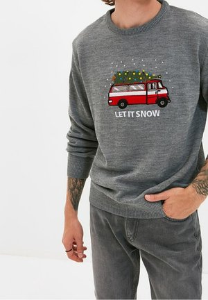 Szary sweter z czerwonym vanem przewożącym udekorowaną choinkę i napisem „LET IT SNOW”, noszony przez osobę z tatuażami i szarymi spodniami.