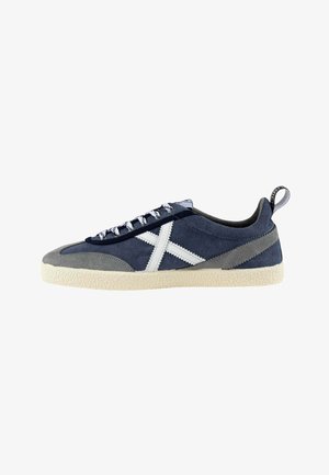 Zapatilla casual de corte bajo en azul y gris con diseño blanco cruzado, suela blanca, cordones con estampado y tira de sujeción negra en el talón.