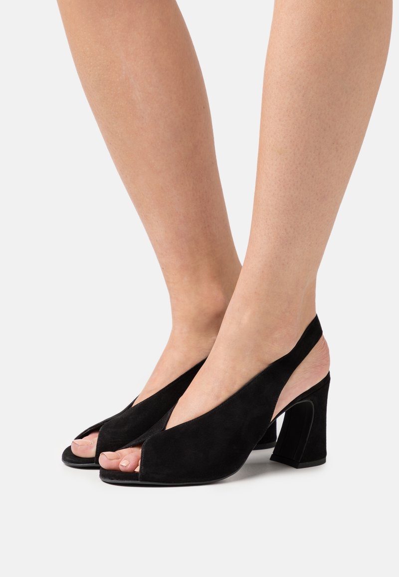 Pieds portant des escarpins slingback en daim noir à bout ouvert avec un talon bloc de hauteur moyenne sur fond blanc.