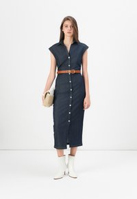 Denim midi-kjole med krage, korte ermer, frontknapper og et brunt belte. Hvite ankelstøvler og en liten vevd håndveske fullfører antrekket.
