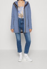 Felpa con cappuccio blu, con zip frontale, maniche lunghe e dettagli grigi, indossata sopra una maglietta a fantasia e abbinata a jeans blu e sneakers bianche.