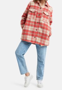 Rot und cremefarbener karierten Flanellhemdjacke mit zwei Brusttaschen und braunen Knöpfen, kombiniert mit hellblauen geraden Jeans und weißen Turnschuhen.