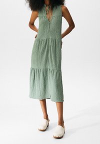 Robe midi texturée vert clair avec un décolleté en V, fermeture à lacer et design à volants. Portée avec des sabots blancs en bois.