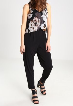 Vrouw met een mouwloze zwarte top met grote witte bloemenprint, losse zwarte broek en zwarte sandalen met hakken en gespen, staand tegen een witte achtergrond.