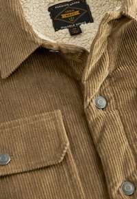 Braunes Cordhemd mit einem fleecegefütterten Kragen, ausgestattet mit silbernen Knöpfen und einer Brusttasche. Strukturiertes Gewebe mit vertikalen Streifen.