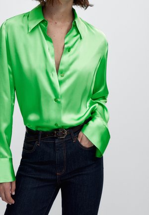 Person trägt ein hellgrünes, satiniertes Hemd mit Knöpfen, das in eine dunkelblaue Jeans mit schwarzem Gürtel gesteckt ist, eine Hand in der Tasche.
