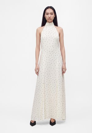 Vrouw draagt een mouwloze witte maxi-jurk met zwarte polkadots en zwarte pumps met puntige neus, staand tegen een effen achtergrond.