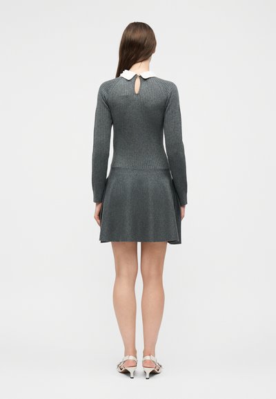 Femme portant une robe longue à manches longues gris côtelée avec un col blanc et un détail œillet dans le dos, assortie à des sandales blanches à talons, debout de dos.