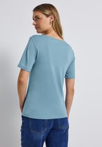 Ljusblå kortärmad t-shirt med rund halsringning. Slät yta, figurnära design. Bärs med mörkblå jeans. Bakvy.