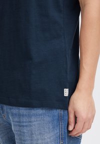 Námornícky modré tričko s jemnou textúrou, so malou látkovou etiketou na okraji. V kombinácii so svetlomodrými džínsami.