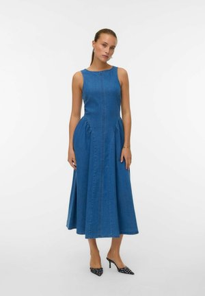 Robe midi en denim avec un corsage ajusté, une jupe évasée et des plis sur les côtés. Tissu bleu avec des détails de couture visibles. Talons à pois noirs.