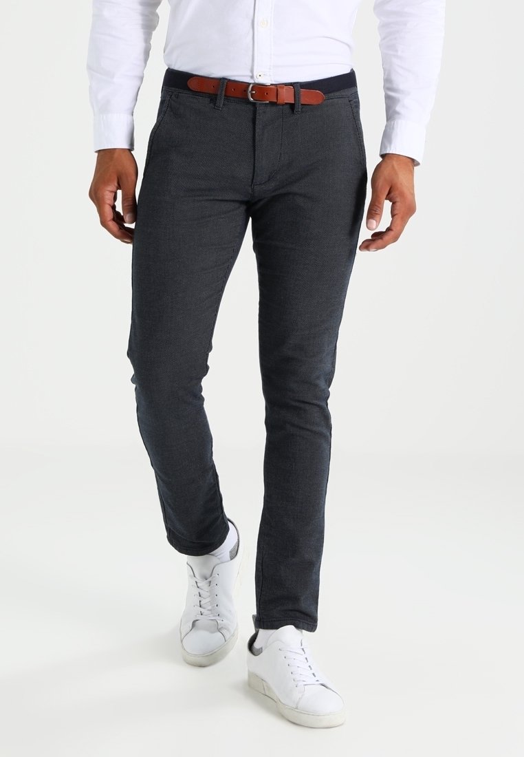 Pantalon bleu marine sur mesure en tissu texturé, coupe slim, associé à une ceinture marron et des baskets blanches, présentant une surface lisse et des lignes épurées.