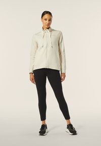 Felpa zip con colletto alto di colore crema, dettagli con lacci e orlo arrotondato, abbinata a leggings neri e scarpe da ginnastica nere.