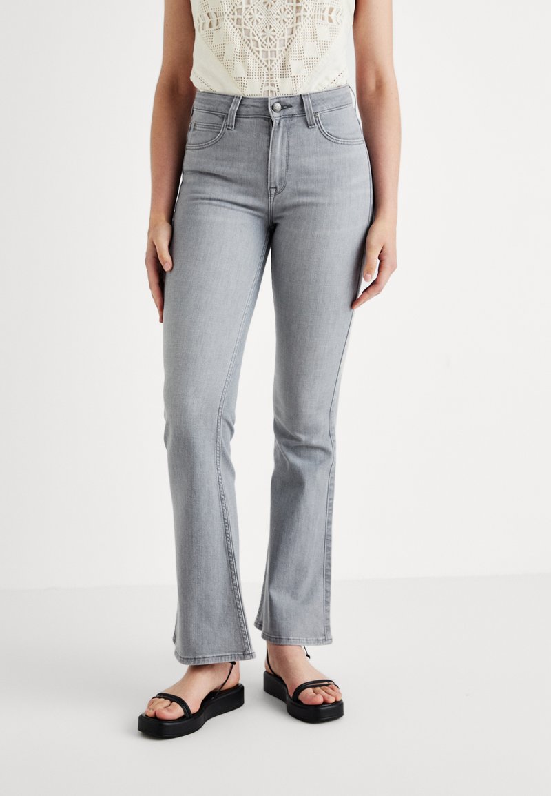 Jeans a zampa d'elefante in denim grigio chiaro con vita alta, tasche frontali e una leggera finitura sbiadita, abbinati a sandali neri flat.