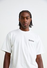 Hvit bomull t-skjorte med korte ermer, med en liten svart "Dickies" logo på venstre side av brystet. Myk tekstur og avslappet passform.