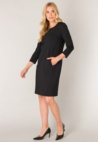 Zwarte jurk met driekwart mouwen, ronde halslijn en zijzakken. Gemaakt van gladde stof met een aansluitende silhouet. Draagbaar met zwarte hakken.