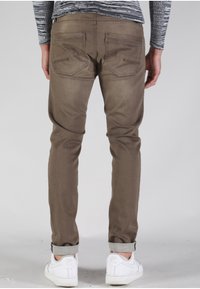 Vaqueros slim-fit marrones con bolsillos traseros, puños doblados y detalles de costura sutiles, combinados con zapatillas blancas.