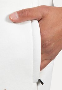 Tejido de sudadera blanco con una mano en un bolsillo. El bolsillo tiene una pequeña pestaña negra y una textura acanalada a lo largo de los bordes.