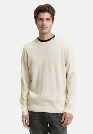 Pull léger beige en maille douce et texturée, avec un col rond et des poignets à côtes, porté par-dessus un t-shirt noir.