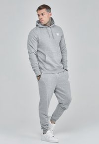 Light gray hoodie with a front pocket and white logo, paired with matching gray sweatpants, and white sneakers. Smooth fabric texture.