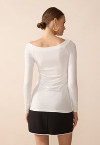 Bijela bluza dugih rukava s off-shoulder izrezom, mekanog tekstila i naglašenim naborima na leđima, u kombinaciji s crnom suknjom.