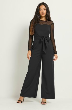 Vrouw draagt een zwarte jumpsuit met doorzichtige lange mouwen, een gestrikte tailleceintuur en brede pijpen, staand met één hand ontspannen langs haar zij.