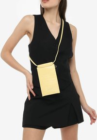 Gul faux læder crossbody taske med et struktureret krokodillemønster, rektangulær form og justerbar rem, båret over et sort outfit.