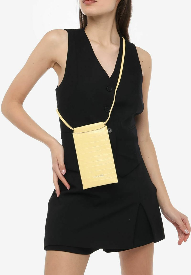 Gul faux læder crossbody taske med et struktureret krokodillemønster, rektangulær form og justerbar rem, båret over et sort outfit.