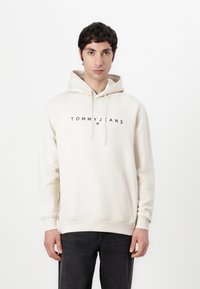 Crèmekleurige hoodie met een voorzak, capuchon met trekkoord en het "TOMMY JEANS" logo in het zwart over de borst. Zachte stof, relaxed fit.