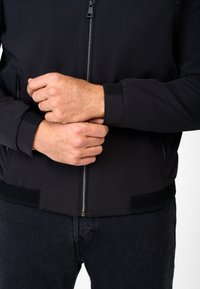Zwarte bomberjack met een gladde, matte afwerking en geribde manchetten. Voorzien van een ritssluiting aan de voorkant en mesh accentueringen aan de zoom. Donkere spijkerbroek zichtbaar eronder.