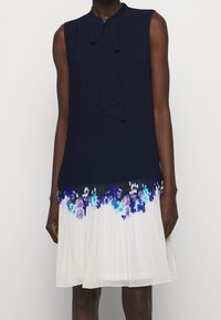 Robe sans manches bleu marine à texture côtelée, avec un col noué en forme de lavallière et une jupe plissée blanche ornée d'accents floraux bleus et violets.