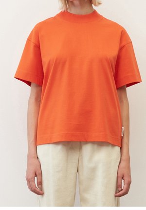 Person trägt ein oranges Kurzarm-T-Shirt und cremefarbene Cordhosen, steht vor einem schlichten hellen Hintergrund.