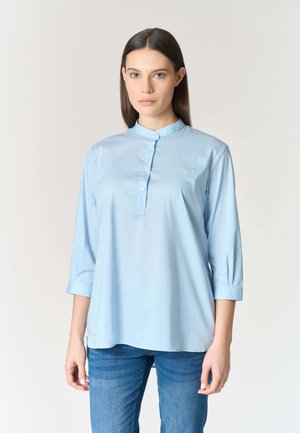 Donna con capelli castani lisci indossa una camicetta azzurra con bottoni e maniche a tre quarti, abbinata a jeans blu, in piedi davanti a uno sfondo semplice.