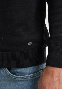Schwarzer Strickpullover mit geripptem Saum und Bündchen. Mit kleinem silbernen Logo-Detail. Kombiniert mit blauer Jeans, die eine Tasche zeigt.
